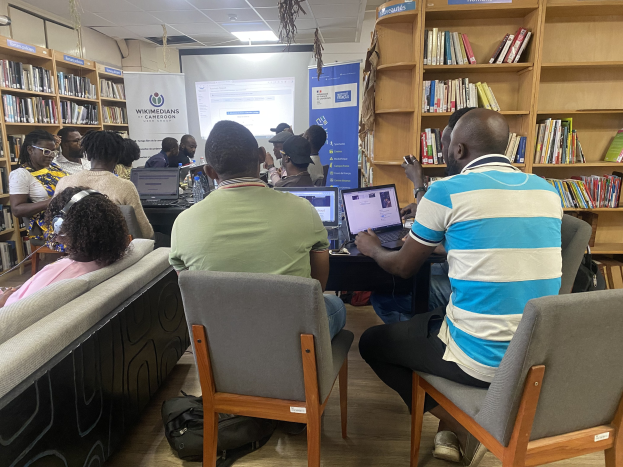 Gruppe von Menschen an einem Tisch mit Laptops in einer Bibliothek, umgeben von Bücherregalen, mit einem Banner, Bildschirm, Deckenleuchten und Taschen auf dem Boden.