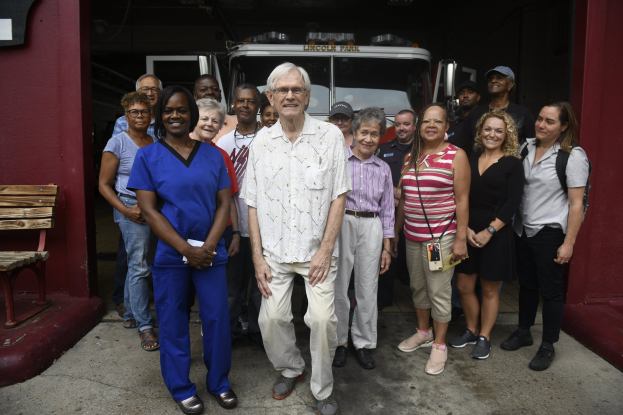 Eine Gruppe von Freiwilligen vor einem Feuerwehrfahrzeug, mit einer Bank links und einer Wand im Hintergrund.