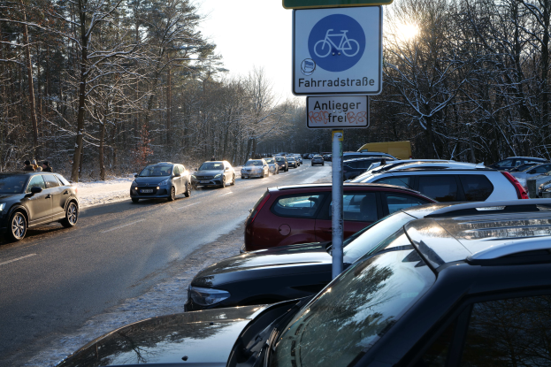 Ein Stau auf einer verschneiten Straße mit parkenden Autos am Rändern, ein Verkehrsschild im Vordergrund und Tannen im Hintergrund.