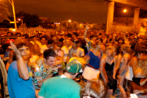 Leute tanzen im Vordergrund bei einem Konzert, mit Lichtern, Pfählen, Bäumen und anderen Gegenständen im Hintergrund.