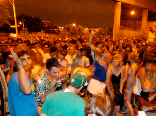 Leute tanzen im Vordergrund bei einem Konzert, mit Lichtern, Pfählen, Bäumen und anderen Gegenständen im Hintergrund.