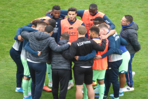 Eine Gruppe von Männern in Jacken steht zusammen auf einem Füßballfeld.