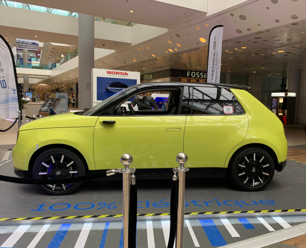 Ein gelber wasserstoffbetriebenes Elektroauto vor einem Gebäude mit Absperrpfosten, Bannern, Menschen, Schildern, einer Topfpflanze, Deckenleuchten und Ventilatoren.