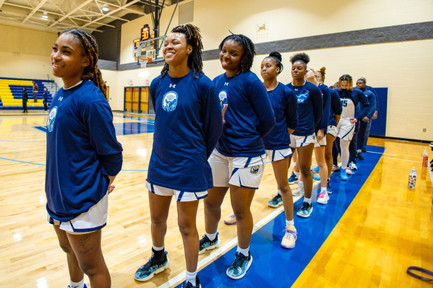 Eine Gruppe junger Frauen in blauen T-Shirts lächelt auf einem Basketballfeld, mit Flaschen und Gegenständen rechts und Zuschauern im Hintergrund.