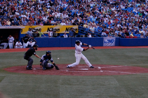 Ein Mann h"alt einen Baseballschl"ager mit einem Ball hinter sich, w"ahrend mehrere Personen auf St"uhlen sitzen und andere Kameras halten im Hintergrund, mit Werbetafeln und Metallst"angen davor.