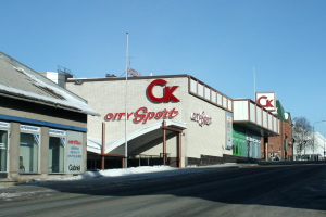 Außenansicht eines Stadt-Sportgeschäfts an einer Straßenecke mit umliegenden Gebäuden, Straßenmöbeln, Bäumen und Schnee unter einem sichtbaren Himmel.