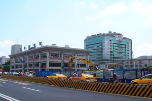Eine belebte Stadtstra├če mit zahlreichen Fahrzeugen, Fußgängern, hohen Gebäuden, einem Kran, Verkehrsampeln, Schildern, Bäumen und einem bewölkten Himmel.