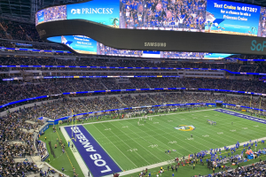 Großes Stadion voller Zuschauer, die ein Fußballspiel auf saftig grünem Rasen verfolgen, mit Spielern auf dem Feld und einem großen Bildschirm mit Text und Bildern oben.