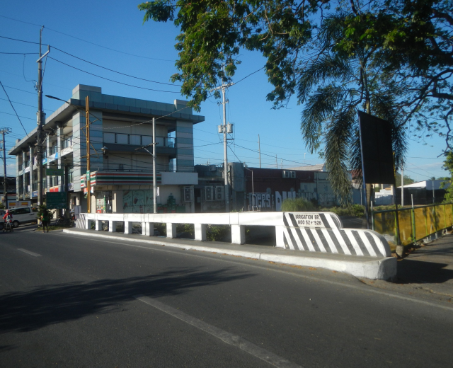 Stadtstraße mit Gebäuden, Strommasten, Kabeln, Fahrzeugen, Absperrpollern, Schildern, Bäumen und einem klaren blauen Himmel.