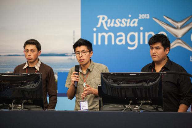 Drei Jungs stehen neben einem Schreibtisch mit zwei Computern, einer spricht in ein Mikrofon, tragen ID-Karten mit roten Markierungen, vor einer Wand mit einem Werbeplakat, auf dem "Russia Imagine 2013" steht.