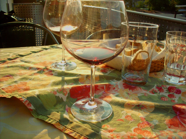 Ein Tisch mit Wein- und Wassergläsern, einem Brotkorb, einer floralen Tischdecke und Stühlen sowie Bäumen im Hintergrund.