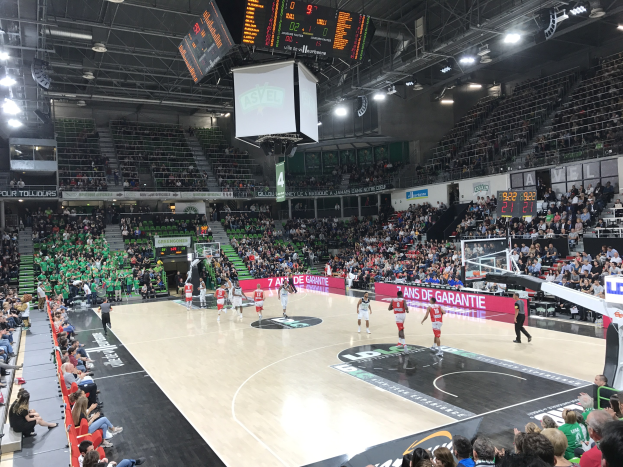 Basketballspiel in einer großen Arena mit Zuschauern, Anzeigetafeln mit "Asvel vs Galatasaray" und Decke mit Lampen und Bildschirm.