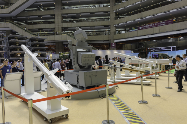Eine Gruppe von Menschen steht in einem großen Raum voller verschiedener Raketen, einige halten Kameras, mit Stangen mit Bändern, Texttafeln, Deckenlampen und anderen Gegenständen im Hintergrund auf der Hong Kong International Defence Exhibition.