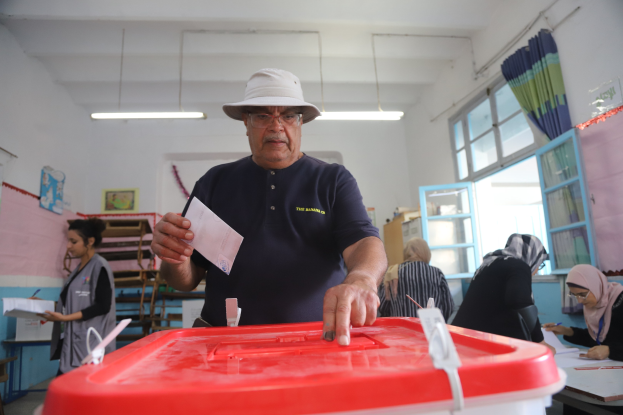 Ein Mann mit Hut stimmt an einer Wahllokal ab, vor einem roten Wahlurne mit einem Stück Papier in der Hand.