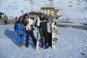 Vier Personen posieren für ein Foto mit Skating-Pads und Werkzeugen, im Hintergrund sind Gebäude und andere Menschen zu sehen, vor einer schneebedeckten Berglandschaft.