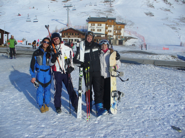 Vier Personen posieren für ein Foto mit Skating-Pads und Werkzeugen, im Hintergrund sind Gebäude und andere Menschen zu sehen, vor einer schneebedeckten Berglandschaft.