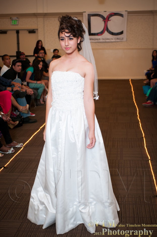 Eine Frau in einem weißen Kleid lächelt und posiert für ein Foto vor einem weißen Banner mit Text, während Menschen auf Stühlen auf beiden Seiten von ihr sitzen und ein brauner Boden unten sichtbar ist.