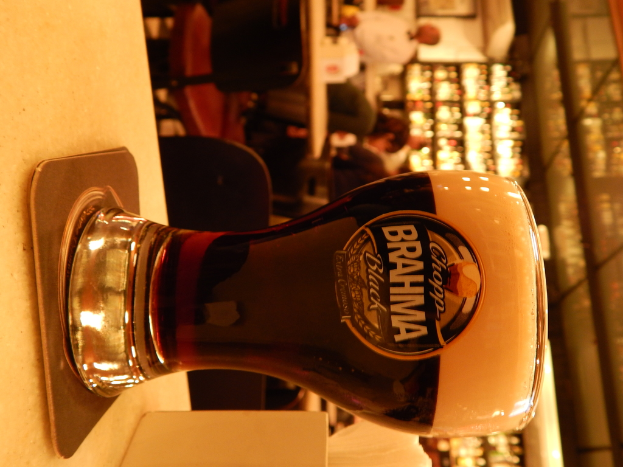 Ein Glas Wein auf einem Teller und eine Papiertaschentuchschachtel auf einem Pfad, mit einer Gruppe von Menschen, Stühlen, Tischen, einem wandmontierten Rahmen und Lichtern im Hintergrund.