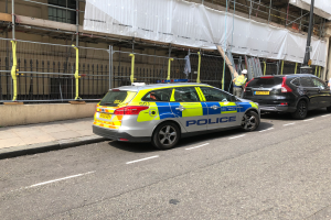 Ein Polizeiwagen steht neben einem Gebäude auf der Straße, daneben stehen zwei Helme tragende Personen, im Vordergrund eine Reling und ein Pfahl, im Hintergrund ein weißes Tuch über das Gebäude drapiert, was auf eine polizeiliche Untersuchung nach einem Messerangriff in London hinweist.