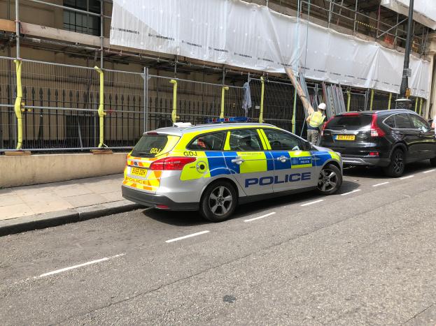 Ein Polizeiwagen steht neben einem Gebäude auf der Straße, daneben stehen zwei Helme tragende Personen, im Vordergrund eine Reling und ein Pfahl, im Hintergrund ein weißes Tuch über das Gebäude drapiert, was auf eine polizeiliche Untersuchung nach einem Messerangriff in London hinweist.
