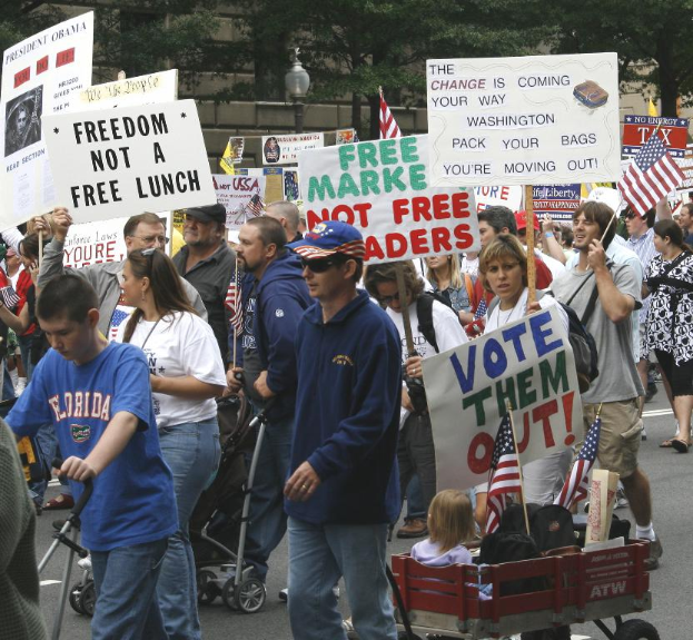 Eine Gruppe von Menschen, die auf einer Straße gehen und Schilder tragen, mit Kindern in einem Wagen vorne und einem Baum, einem Laternenpfahl und einem Gebäude im Hintergrund.