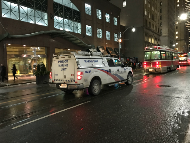 Ein Polizei-Lkw parkt nachts am Straßenrand, mit Fahrzeugen auf der Straße, Fußgängern auf dem Gehweg, Topfpflanzen, Straßenlaternen, Gebäuden mit Fenstern und einer bewölkten Himmel.
