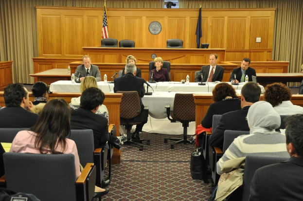 Ein Tagungsraum mit einer Gruppe von Menschen, die auf St├╝hlen sitzen, zwei fremde Flaggen, ein oranger Schreibtisch mit Wasserflaschen und ein Mikrofon.