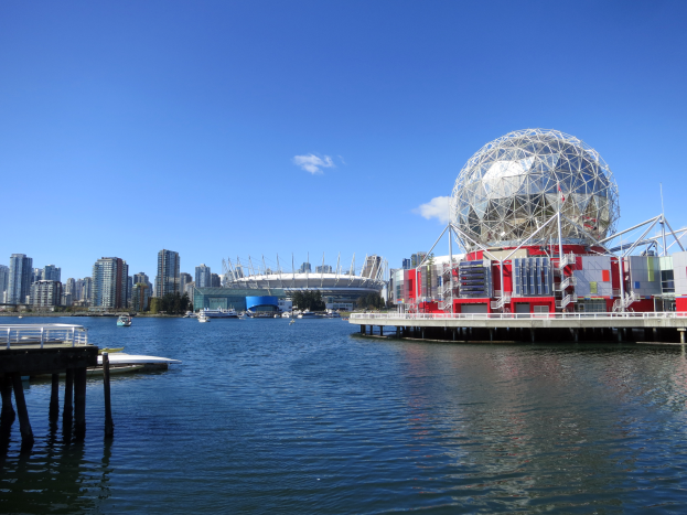Ein großes kugelförmiges Gebäude, Science World, das auf dem Wasser in Vancouver steht, mit einer Brücke auf der linken Seite, Booten auf der Oberfläche und Gebäuden, Bäumen und einem klaren blauen Himmel im Hintergrund.