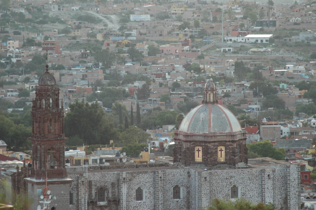 Eine Luftaufnahme einer Stadt mit Bäumen, Gebäuden, Straßen, Masten und einer Kirche im Vordergrund.