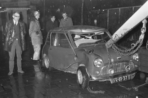 Ein Schwarz-Weiß-Bild eines Wagens, der von einem Abschleppwagen gezogen wird, umgeben von mehreren Menschen, einige mit Helmen, und einem Gebäude im Hintergrund.