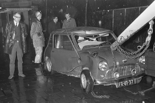 Ein Schwarz-Weiß-Bild eines Wagens, der von einem Abschleppwagen gezogen wird, umgeben von mehreren Menschen, einige mit Helmen, und einem Gebäude im Hintergrund.