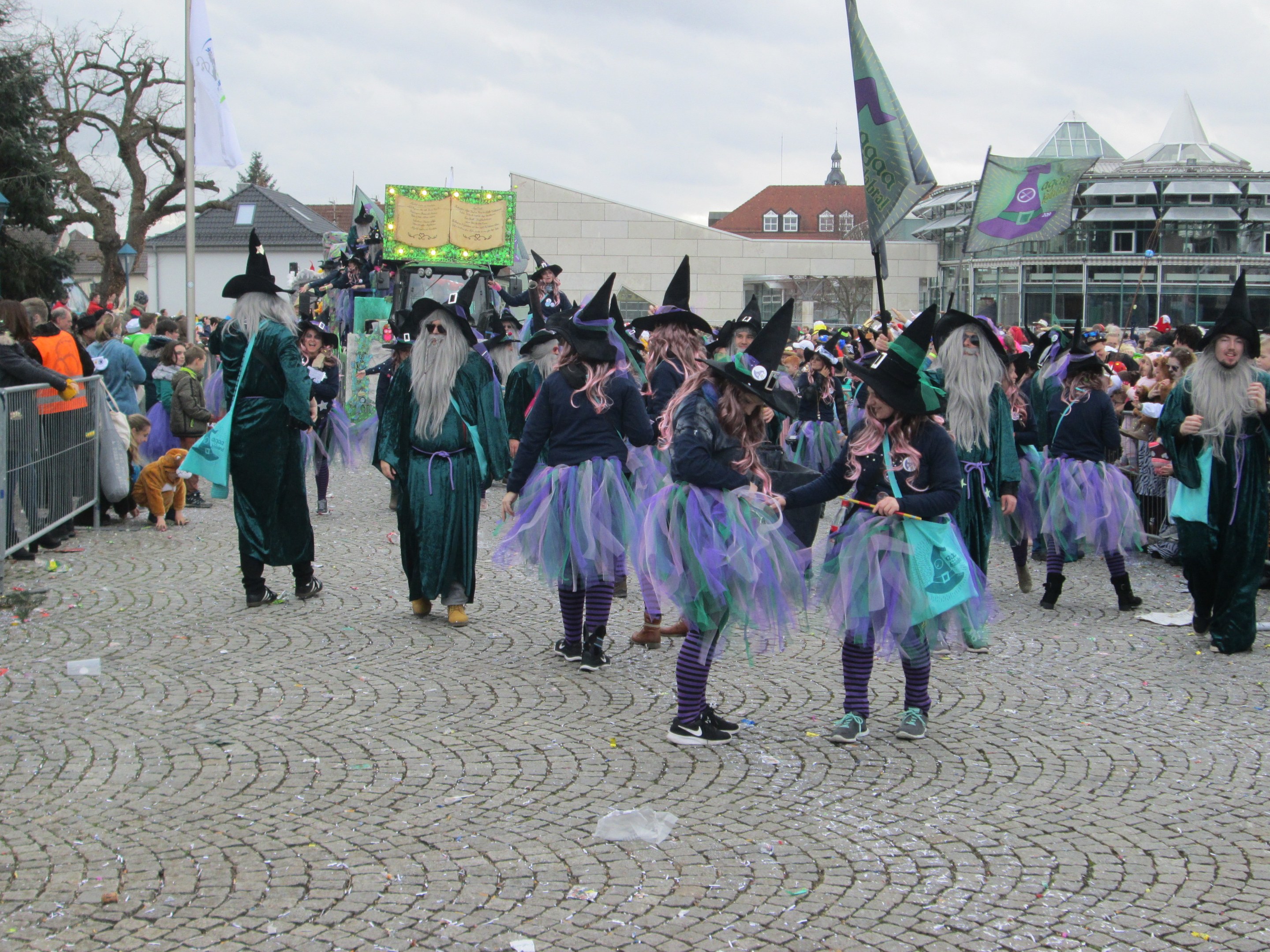 Eine Gruppe von Menschen in Hexenkostümen, die schwarze und lila Kleider und Hüte tragen und einige Fahnen halten, gehen eine Kopfsteinpflasterstraße entlang, neben einem Geländer und vor einer Kulisse aus Gebäuden, Bäumen und einem klaren blauen Himmel.