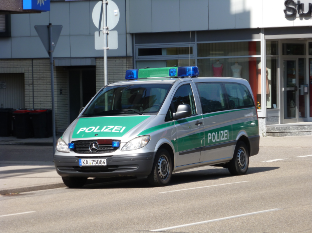 Ein Polizeiwagen parkt am Straßenrand vor einem Gebäude mit Glastüren, die Puppen zeigen, mit Schildern und Mülltonnen im Hintergrund.