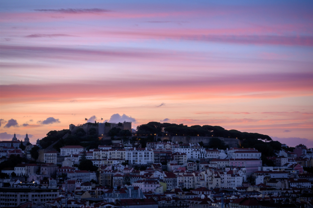 Ein Blick auf Lissabon, Portugal bei Sonnenuntergang von einem Hügel aus, mit Gebäuden und Bäumen im Vordergrund und Wolken am Himmel.