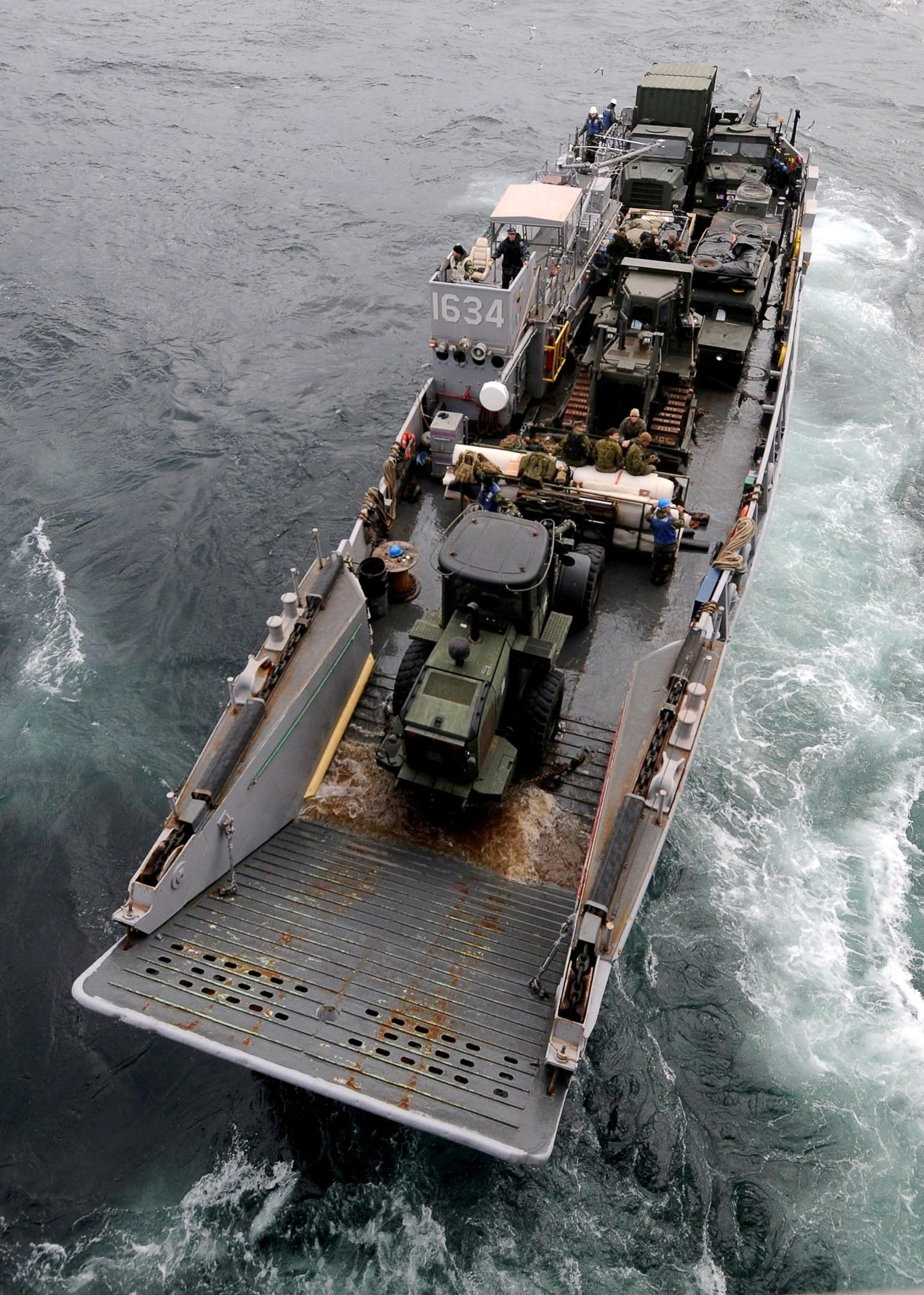 Luftaufnahme eines großen Bootes auf dem Meer, das ein militärisches Fahrzeug trägt, umgeben von Menschen und anderen Gegenständen, mit einem Wasserzeichen unten.