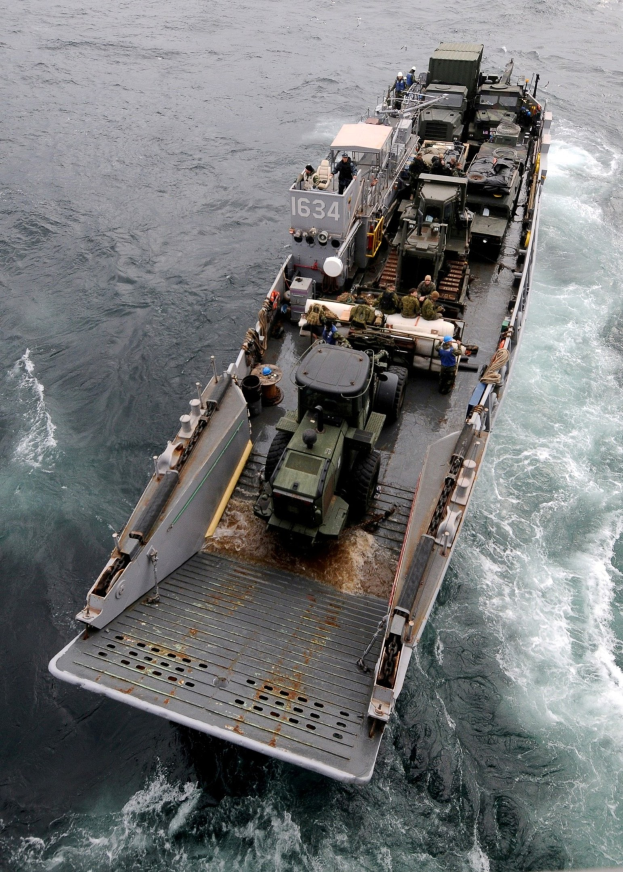 Luftaufnahme eines großen Bootes auf dem Meer, das ein militärisches Fahrzeug trägt, umgeben von Menschen und anderen Gegenständen, mit einem Wasserzeichen unten.
