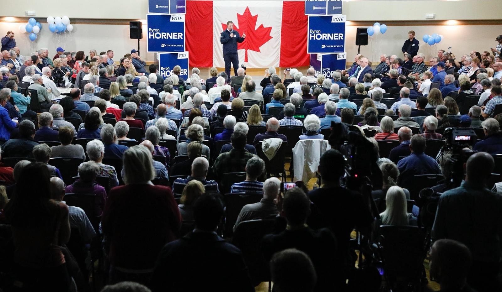 Eine große Menschenmenge sitzt auf Stühlen vor einer kanadischen Flagge, während ein Mann auf einem Podium ein Mikrofon hält, begleitet von Bannern, Luftballons und Lautsprechern.