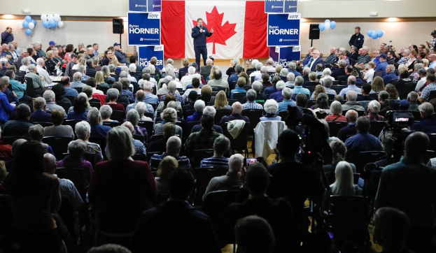 Eine große Menschenmenge sitzt auf Stühlen vor einer kanadischen Flagge, während ein Mann auf einem Podium ein Mikrofon hält, begleitet von Bannern, Luftballons und Lautsprechern.