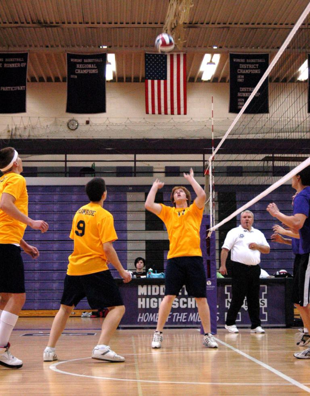 Leute stehen auf einem Spielfeld mit Netz und Ball, tragen Kleidung und Schuhe, mit einem Landesflagge, Wand und Plakaten mit Text im Hintergrund.