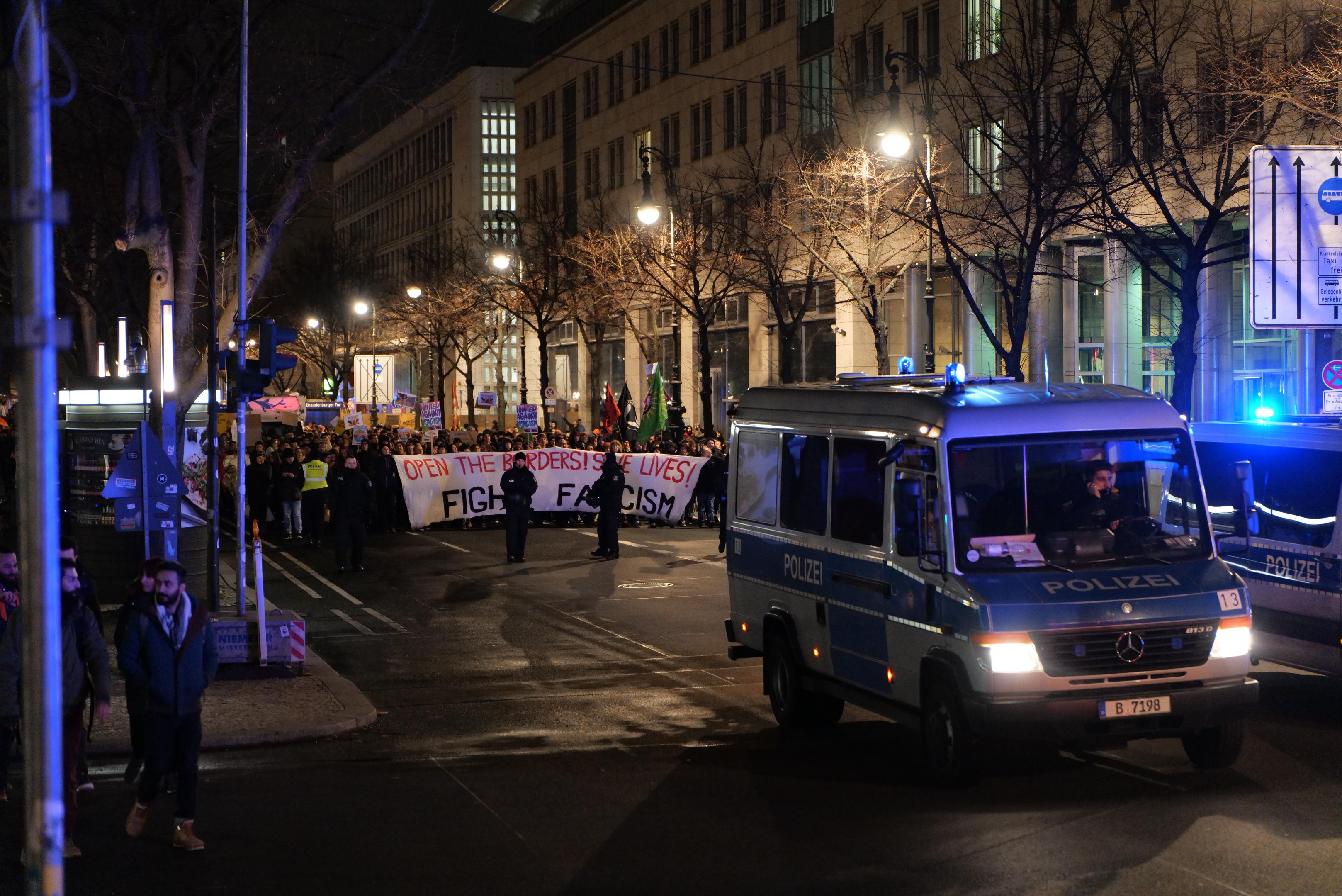 Gruppe von Menschen, die nachts eine von Straßenlaternen beleuchtete Straße entlanggehen, mit einem geparkten Polizeiwagen am Straßenrand und einem Banner im Hintergrund.
