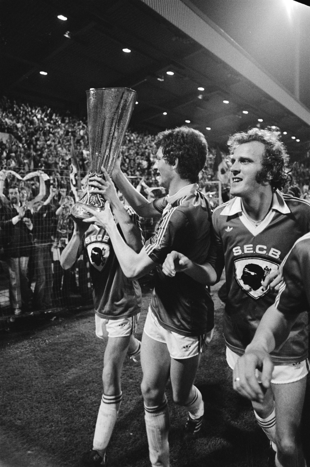 Schwarzes und weißes Foto von Fußballspielern, die einen Pokal auf einem Feld halten und feiern, während Zuschauer hinter einem Zaun unter Stadionlichtern zuschauen.