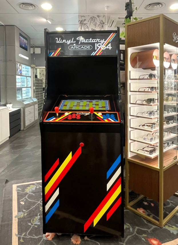 Ein Geschäft mit einem vintage Arcade-Automaten in der Mitte, einer Glasvitrine auf der rechten Seite und Retro-Beleuchtung mit Schränken und einem Fernseher im Hintergrund.
