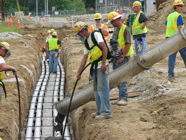 Eine Baustelle mit Arbeitern in Jacken und Helmen, Absperrungen, Zaun, Bäume, Pfosten, Erde und Rohrleitungen im Hintergrund.