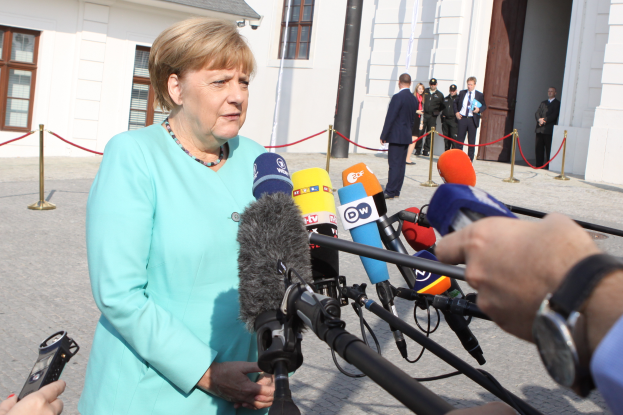 Bundeskanzlerin Angela Merkel spricht vor einem Gebäude mit Fenstern und einer Tür zu den Medien, umgeben von einer Gruppe von Menschen, wobei auf der rechten Seite des Bildes eine Kamera und ein Mobiltelefon zu sehen sind, sowie Pfosten mit daran gebundenen Seilen.