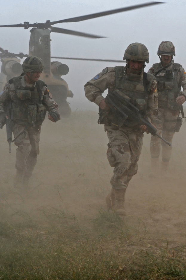 Drei Personen mit Waffen rennen mit einem Hubschrauber im Hintergrund auf einem grasigen und felsigen Boden.