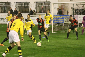 Ein Sportplatz mit mehreren Männern, die Fußball spielen, einem Netz auf der rechten Seite, einer Tafel mit Text im Hintergrund und Zuschauern, die hinter der Tafel stehen, sowie einer Wand und Fenstern weiter im Hintergrund.