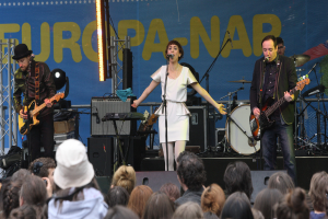 Eine Bühne mit vier Musikern: eine Frau in einem weißen Kleid singt in der Mitte, zwei Männer spielen Gitarren auf beiden Seiten, eine Menge vorne und Lautsprecher im Hintergrund.