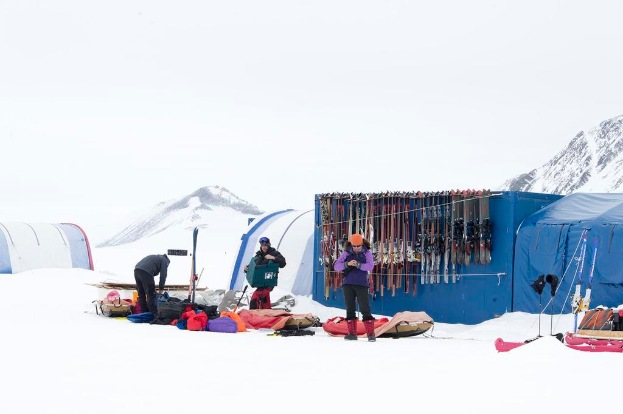 Drei Personen stehen auf einer schneebedeckten Landschaft mit verstreuten Taschen, im Hintergrund Zelte mit Skiern darauf und schneebedeckte Hügel unter einem klaren Himmel.