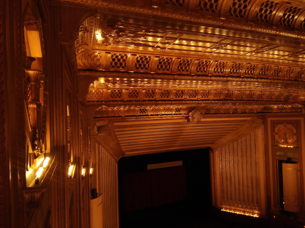 Das Innere eines Theaters mit dunklem Hintergrund, beleuchtet von Lichtern links und rechts, mit einer kunstvoll gestalteten Decke.