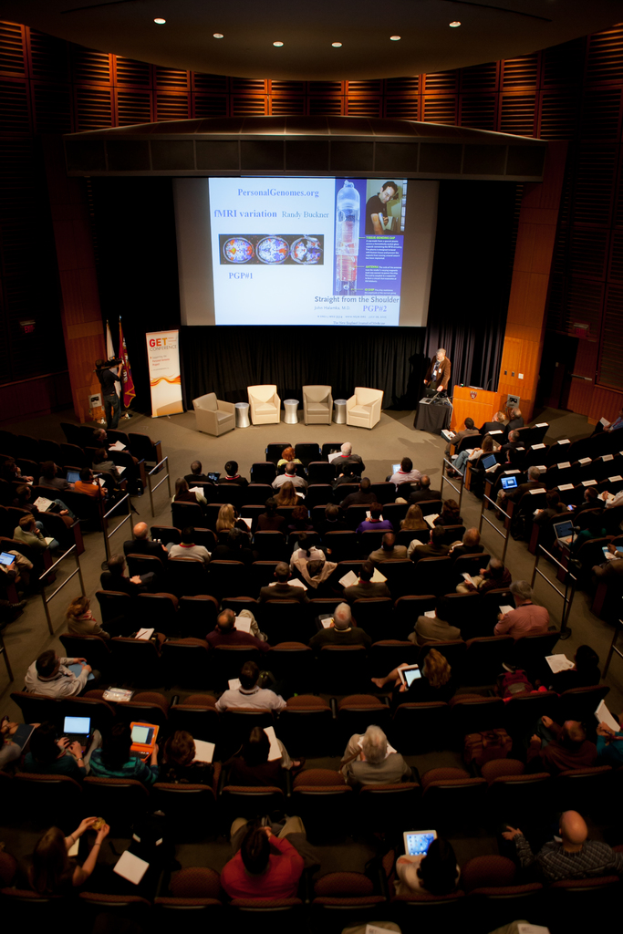 Ein mit Sitzenden und Gegenständen haltenden Menschen gefüllter Hörsaal, beleuchtet von zahlreichen Lichtern, mit Fahnen links, einem Podium rechts und einem Werbeplakat links.